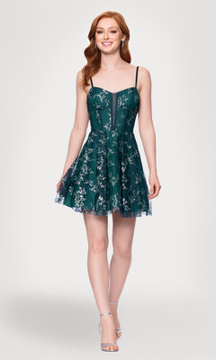 A woman stands and smiles against a plain background, wearing the Short Glitter Hoco Dress: Blondie Nites 4884BN—a sleeveless dark green party dress with floral patterns, spaghetti straps, and silver high-heeled sandals.