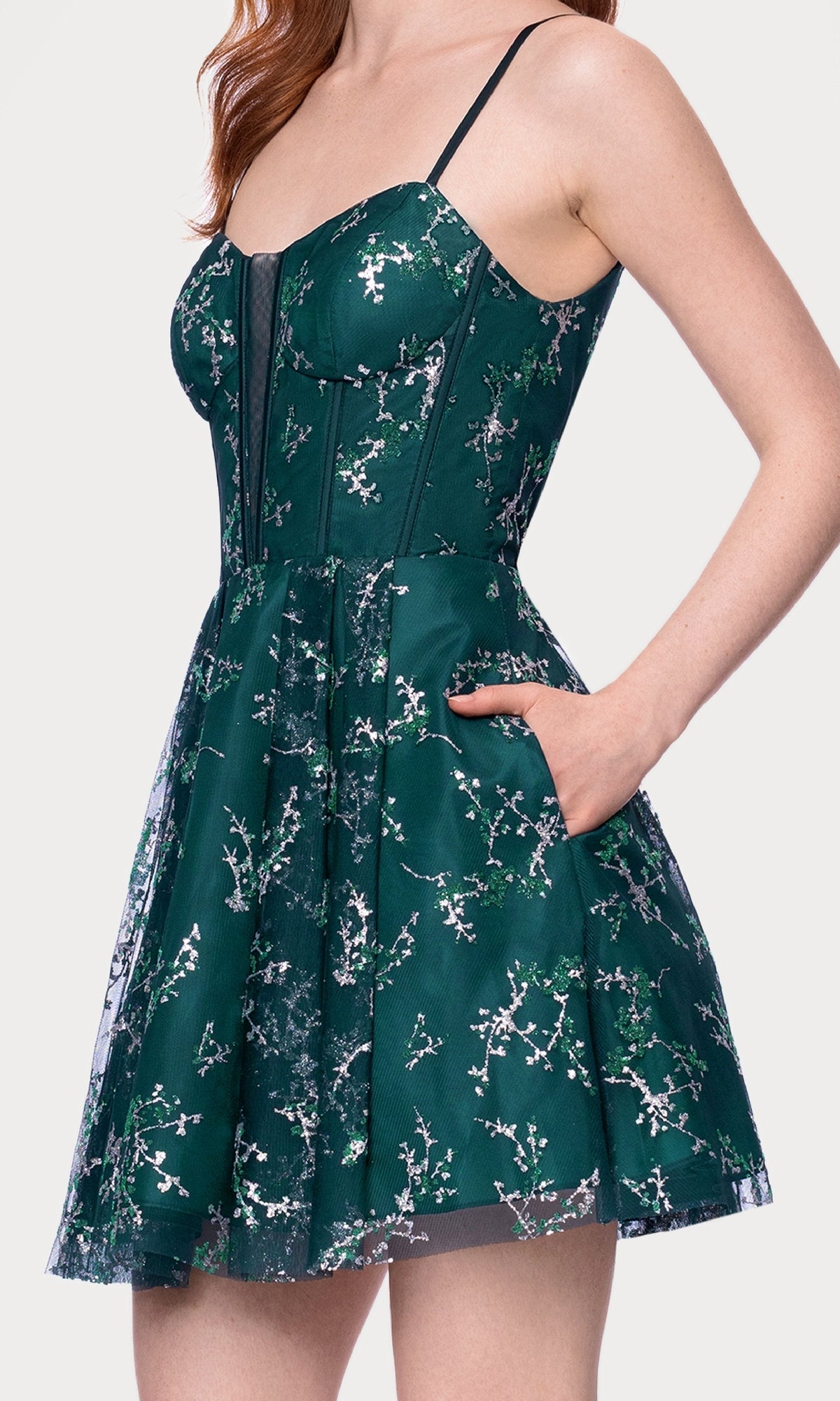 A woman stands against a white background, wearing the Blondie Nites 4884BN short glitter hoco dress—sleeveless with a fitted bodice, flared skirt, and dark green floral pattern.