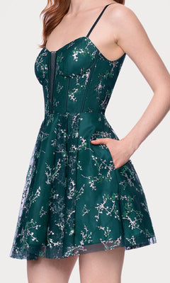 A woman stands against a white background, wearing the Blondie Nites 4884BN short glitter hoco dress—sleeveless with a fitted bodice, flared skirt, and dark green floral pattern.