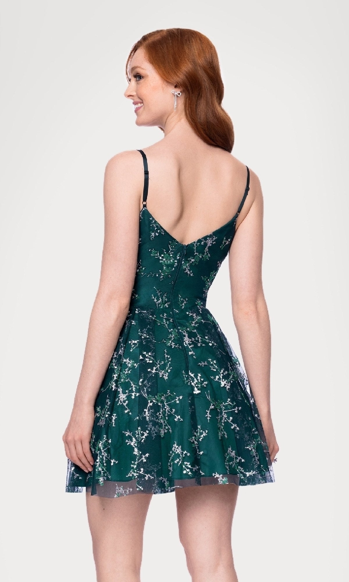 A woman with long red hair wears the Blondie Nites 4884BN short glitter hoco dress, featuring thin straps and a V-shaped back, as she stands smiling over her shoulder.