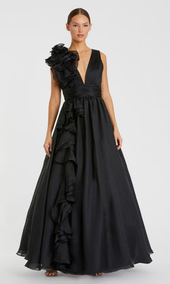 A woman wears the Mac Duggal 48856 Ruffled Long Prom Ball Gown—an elegant black dress with a plunging neckline and ruffled floral detailing on one shoulder and down the front, standing against a plain background.