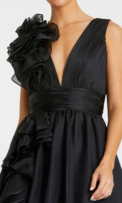 A woman wearing the Mac Duggal 48856 Ruffled Long Prom Ball Gown, featuring an elegant sleeveless black design with a deep V-neckline, pleated bodice, wide waistband, and bold ruffled floral accents—a stunning formal or prom dress.