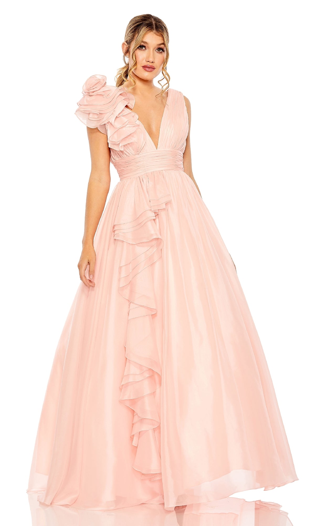A woman wears the Mac Duggal 48856 Ruffled Long Prom Ball Gown, a floor-length light pink dress with a deep V-neck, ruffled details, and a large floral accent on one shoulder.