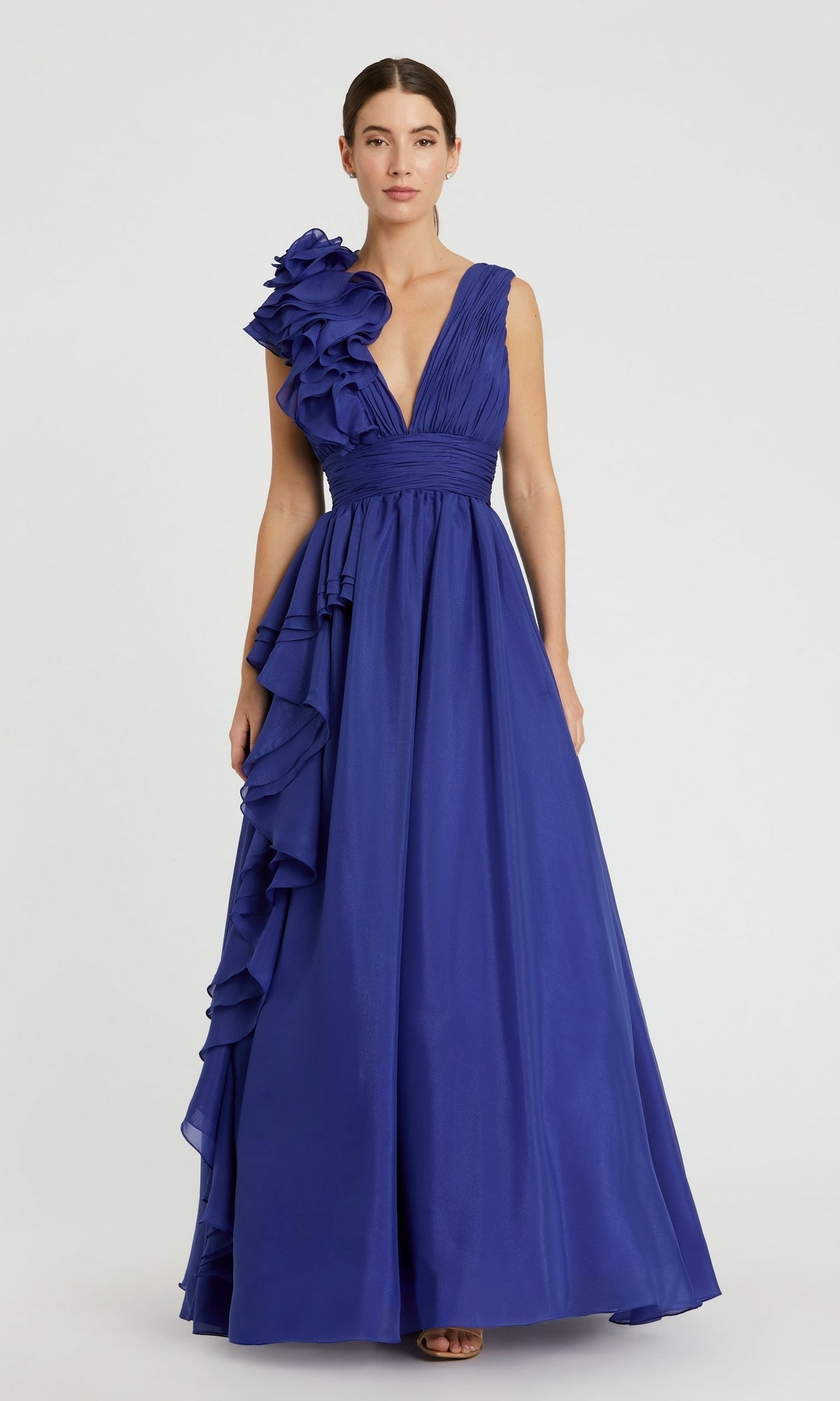 A woman wears the Mac Duggal 48856 Ruffled Long Prom Ball Gown, a sleeveless royal blue A-line evening dress with a deep V-neck and ruffle accents on one shoulder and side.