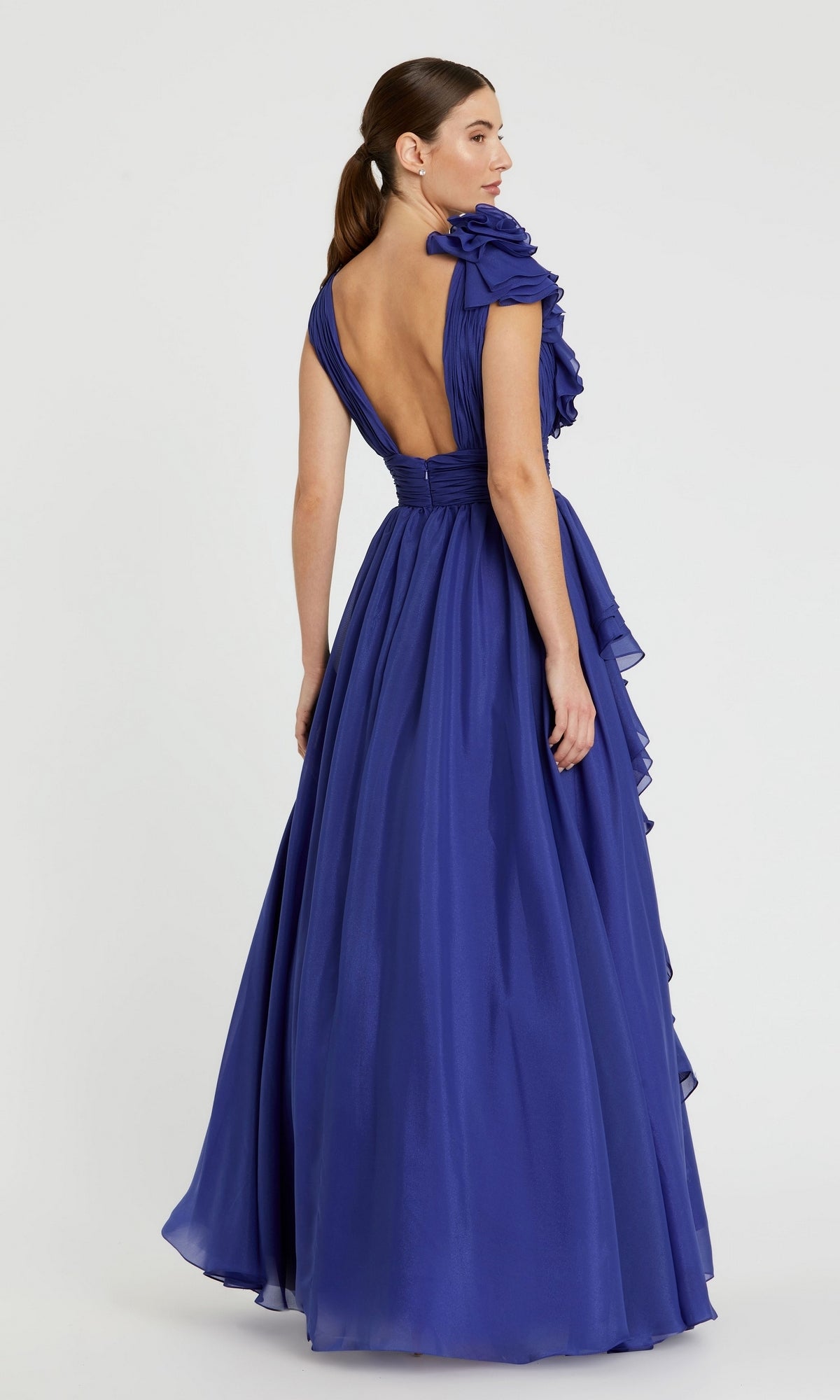A woman models the Mac Duggal 48856 Ruffled Long Prom Ball Gown, featuring a floor-length deep blue design with ruffled shoulder and open back, set against a plain light background.