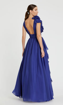A woman models the Mac Duggal 48856 Ruffled Long Prom Ball Gown, featuring a floor-length deep blue design with ruffled shoulder and open back, set against a plain light background.