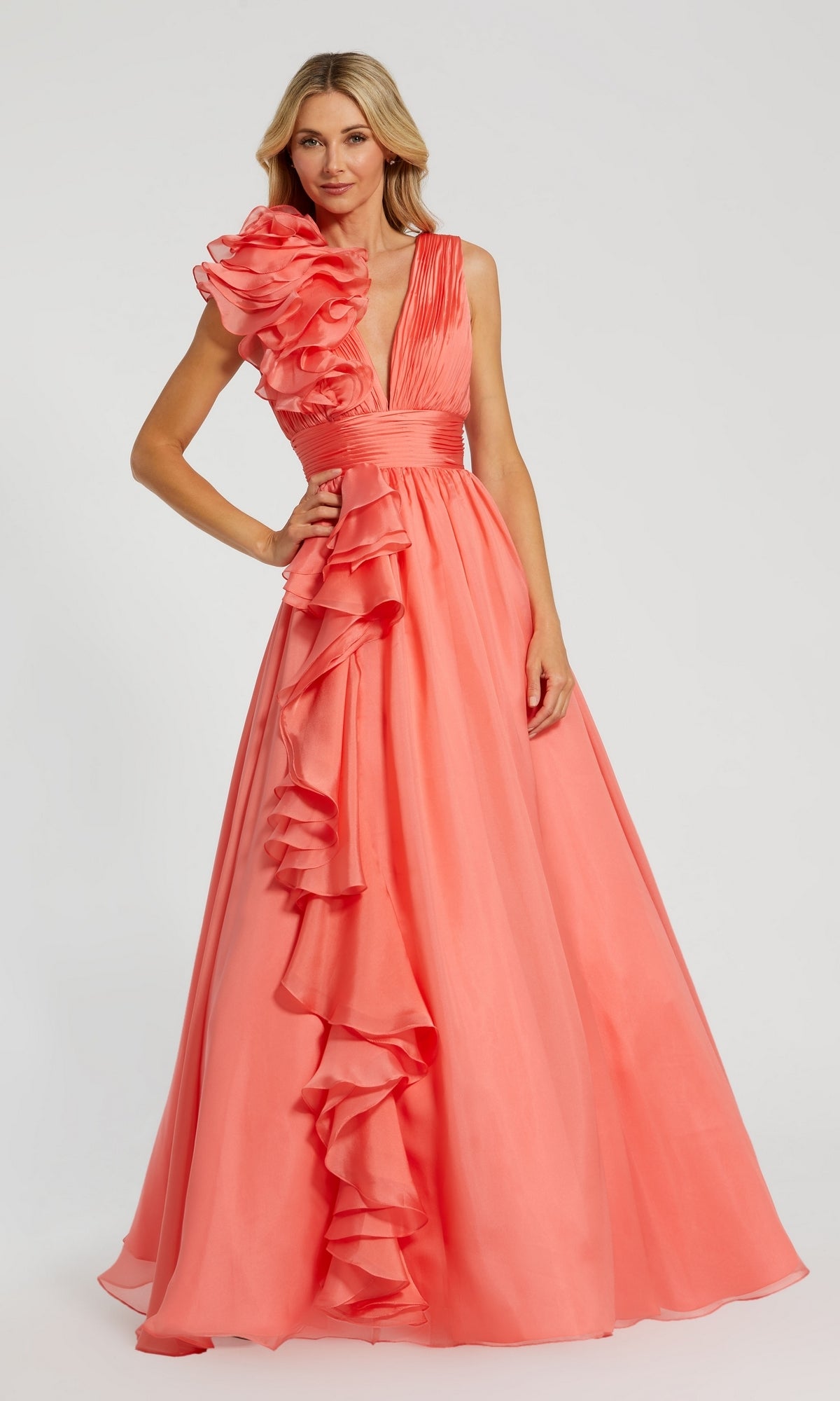 A woman wearing the Mac Duggal 48856 Ruffled Long Prom Ball Gown in coral pink, featuring floor-length style and ruffled details on the bodice and skirt, stands against a plain background.
