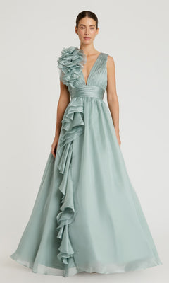 A woman stands against a plain background wearing the Mac Duggal 48856 Ruffled Long Prom Ball Gown in light blue satin, featuring a deep V-neckline, floral ruffle on one shoulder, and cascading skirt ruffles.