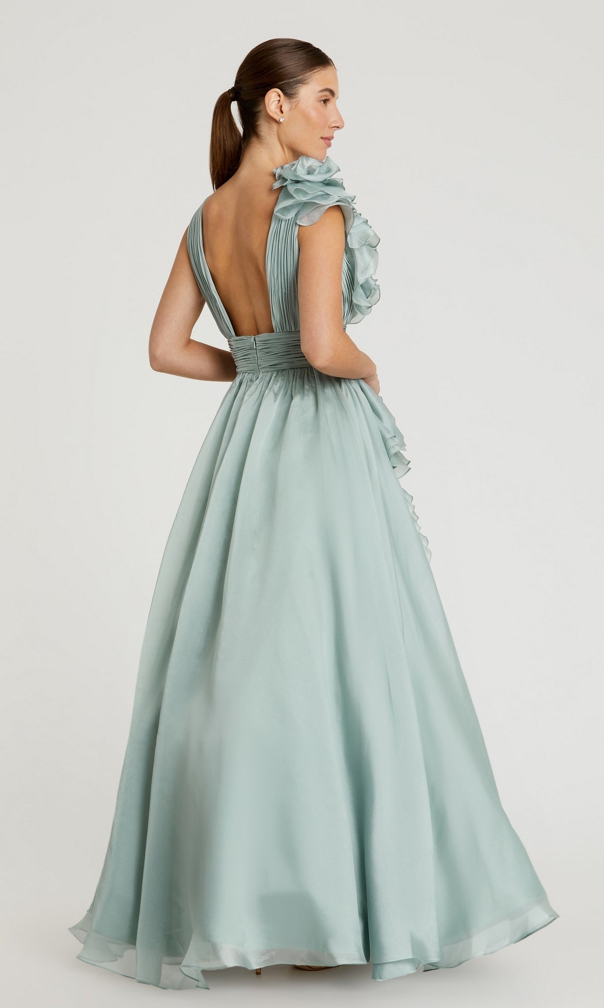 A woman stands against a plain background wearing the Mac Duggal 48856 Ruffled Long Prom Ball Gown, a floor-length light blue dress featuring ruffled shoulder detail and a deep V-back.