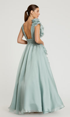 A woman stands against a plain background wearing the Mac Duggal 48856 Ruffled Long Prom Ball Gown, a floor-length light blue dress featuring ruffled shoulder detail and a deep V-back.