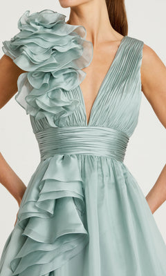 A person wearing the Mac Duggal 48856 Ruffled Long Prom Ball Gown, an elegant light blue A-line dress with pleated details and large ruffled accents on the shoulder and skirt.