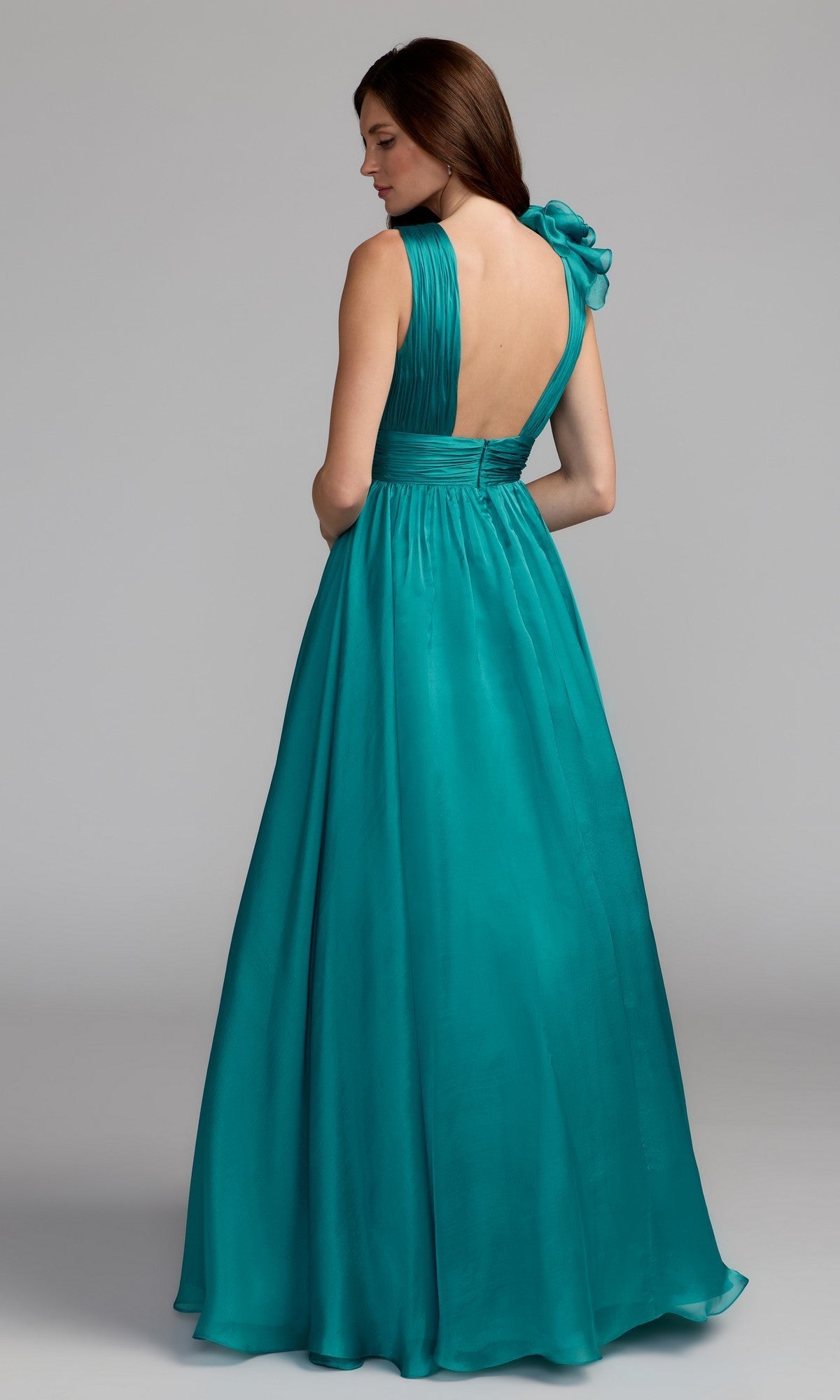 A woman stands with her back turned, wearing the Mac Duggal 48856 Ruffled Long Prom Ball Gown—a sleeveless teal floor-length A-line dress with a single shoulder strap tied in a bow.