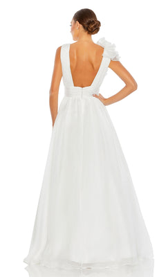 A woman stands facing away in the Mac Duggal 48856 Ruffled Long Prom Ball Gown, featuring a white a-line silhouette, deep V-back, and single ruffled shoulder; her hair is styled in a neat bun.
