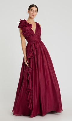 A woman wears the Mac Duggal 48856 Ruffled Long Prom Ball Gown, a floor-length sleeveless burgundy dress with ruffle details on one shoulder and down the side, standing against a plain background.