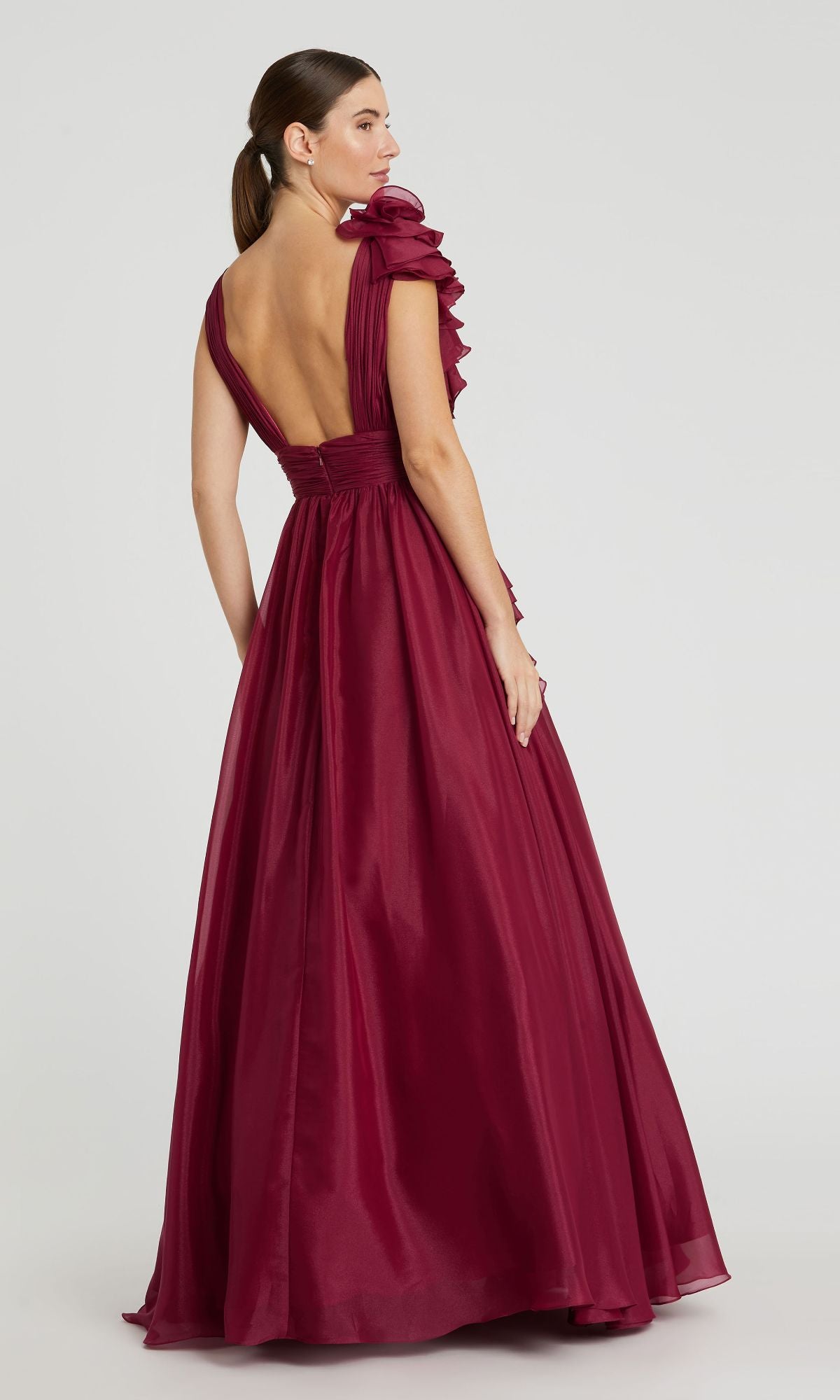 A woman stands against a plain light background wearing the Mac Duggal 48856 Ruffled Long Prom Ball Gown, a floor-length deep red dress with a low back and ruffled shoulder detail.