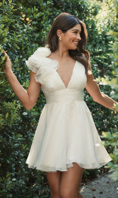 A woman stands outdoors between green bushes, smiling in the Mac Duggal 48952 Ruffle Short White Party Dress with a-line silhouette and ruffled shoulder detail.
