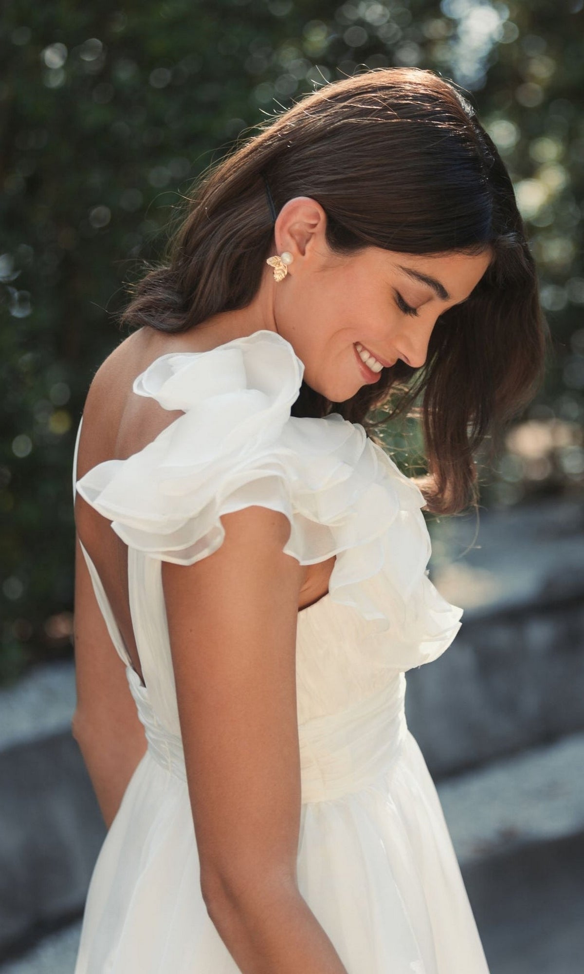 A woman wears the Mac Duggal 48952 Ruffle Short White Party Dress outdoors, smiling and looking down, with greenery behind her.