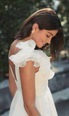A woman wears the Mac Duggal 48952 Ruffle Short White Party Dress outdoors, smiling and looking down, with greenery behind her.