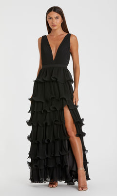 A woman stands against a plain white background wearing the Mac Duggal 49054 V-Neck Long Ruffle Prom Gown, a sleeveless black evening dress featuring a deep v-neckline, layered ruffles, and a high slit.