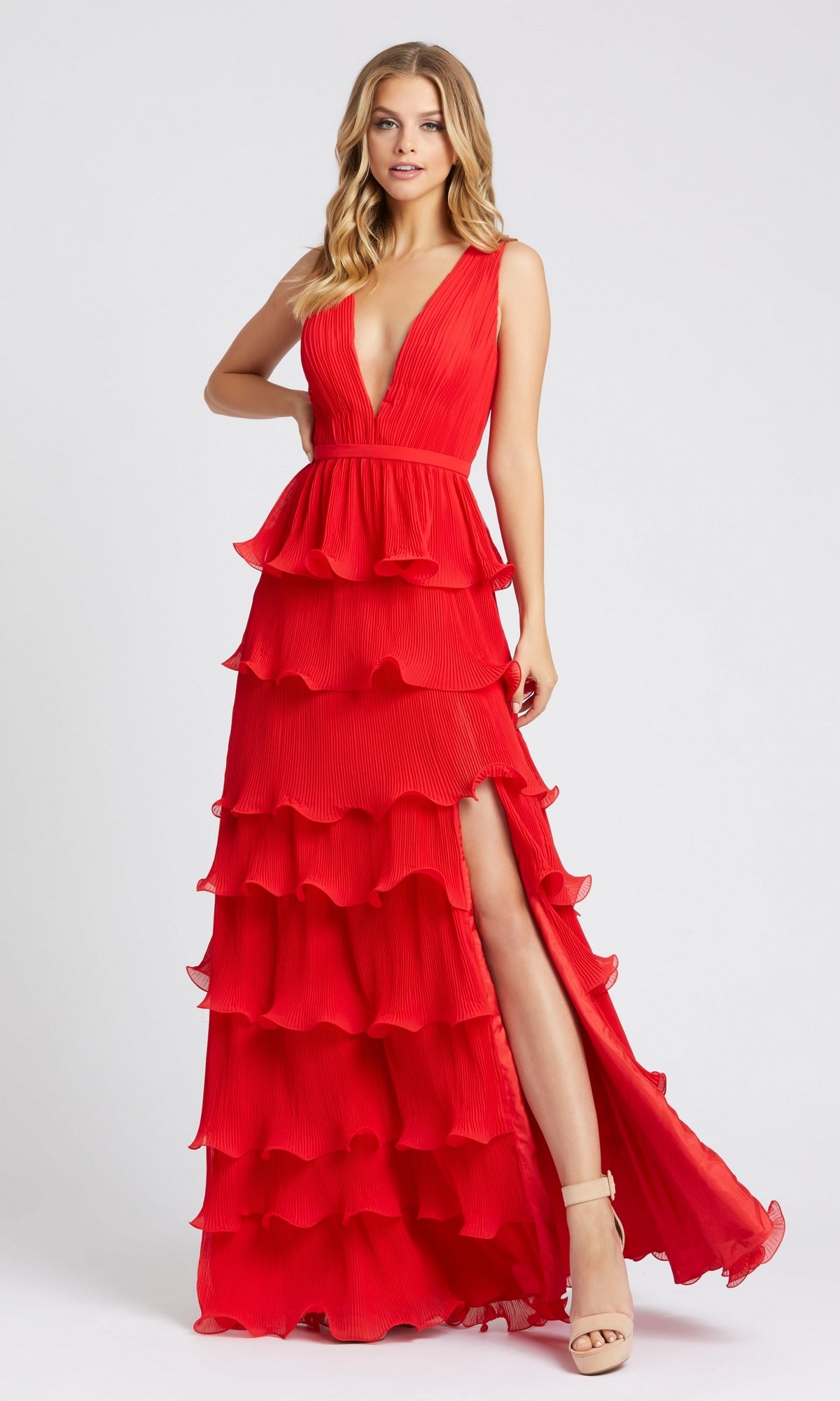 A woman wears the V-Neck Long Ruffle Prom Gown: Mac Duggal 49054, a sleeveless, floor-length dress with tiered ruffles, a deep V-neckline, and high slit. She poses against a plain light background.