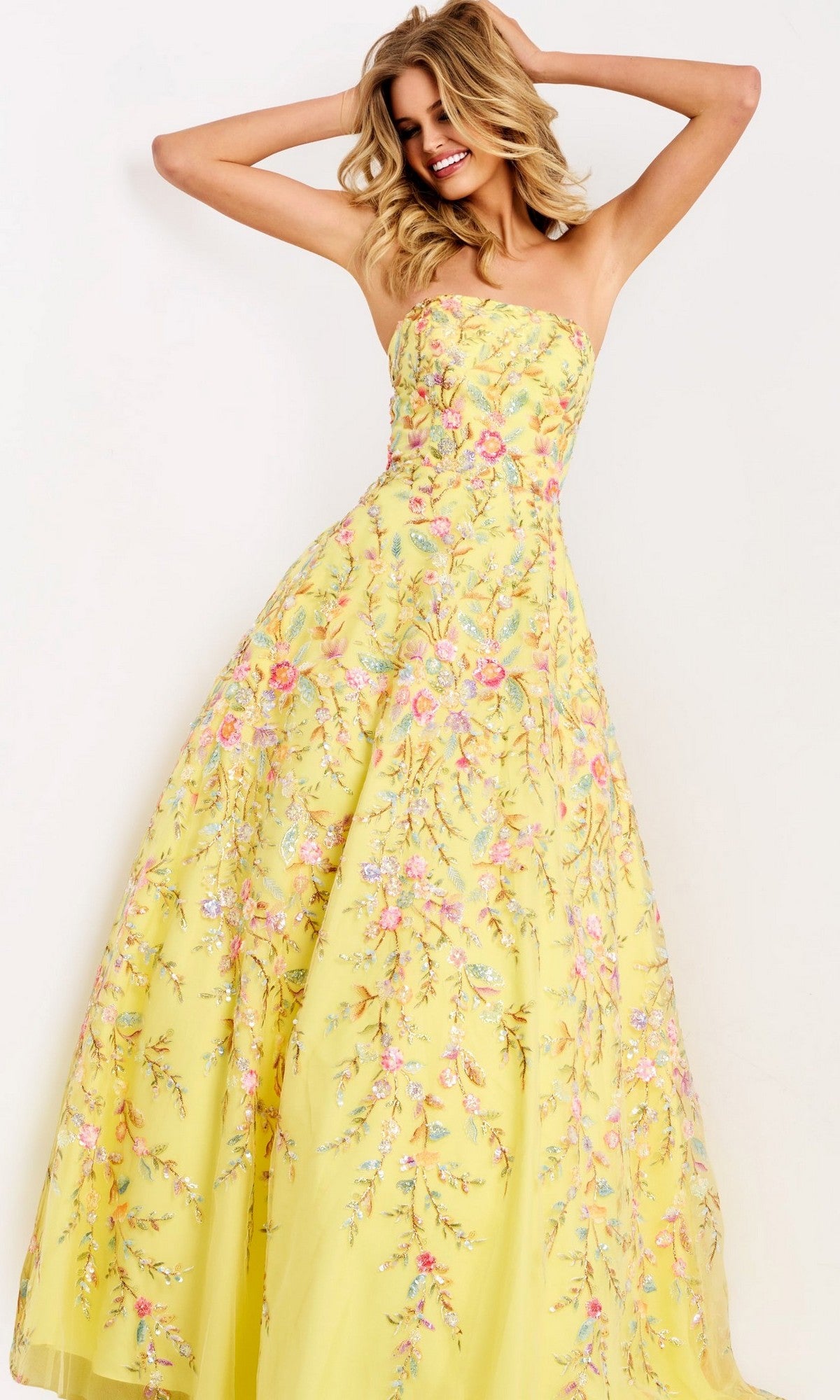 A woman smiles with arms raised, wearing the Jovani 49070— a yellow strapless long prom dress featuring colorful floral embroidery.