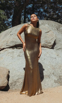 A woman stands outdoors on sandy ground before large rocks and trees, wearing the V-Back Long Metallic Prom Dress: Mac Duggal 49087.