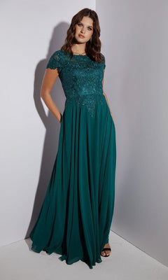 A woman in the Eureka 4909 Short-Sleeve Long A-Line Formal Dress with a teal chiffon skirt and embroidered lace bodice stands against a plain light-colored wall.