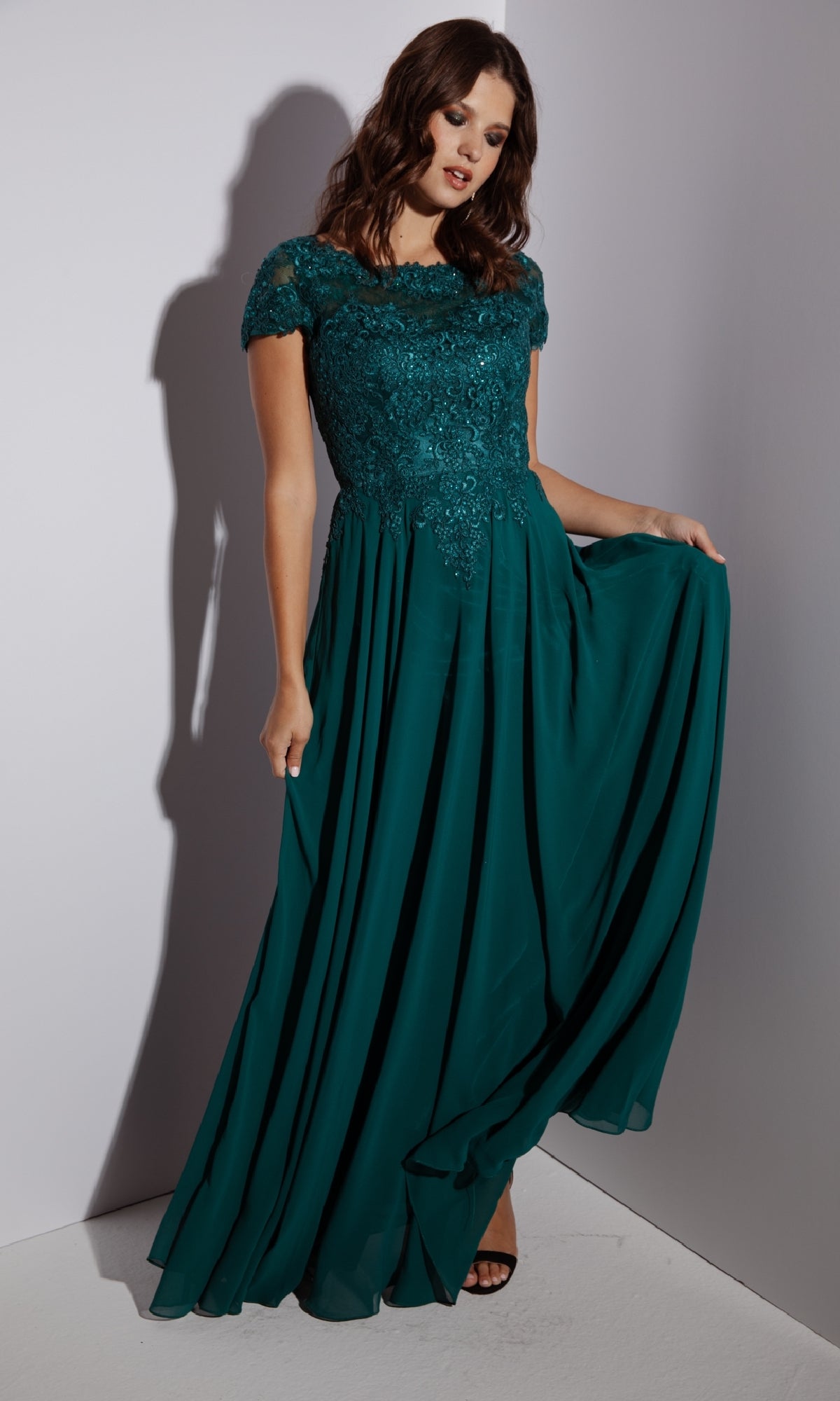Woman in the Eureka 4909 Short-Sleeve Long A-Line Formal Dress, featuring an embroidered lace bodice and flowing chiffon skirt, stands against a plain background while holding the edge of her gown.
