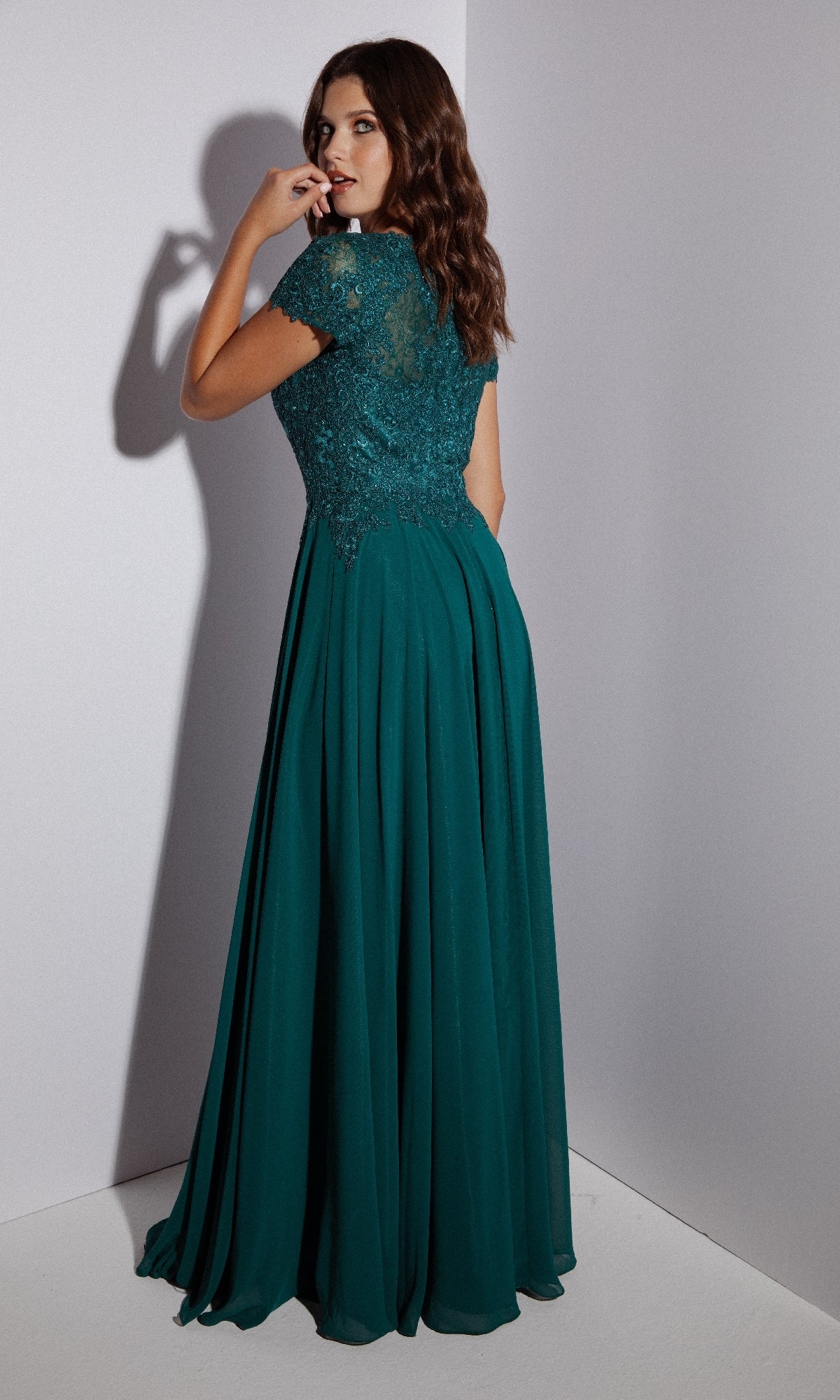Woman with wavy brown hair wears the Eureka 4909 Short-Sleeve Long A-Line Formal Dress with an embroidered lace bodice, standing in a corner and glancing over her shoulder.