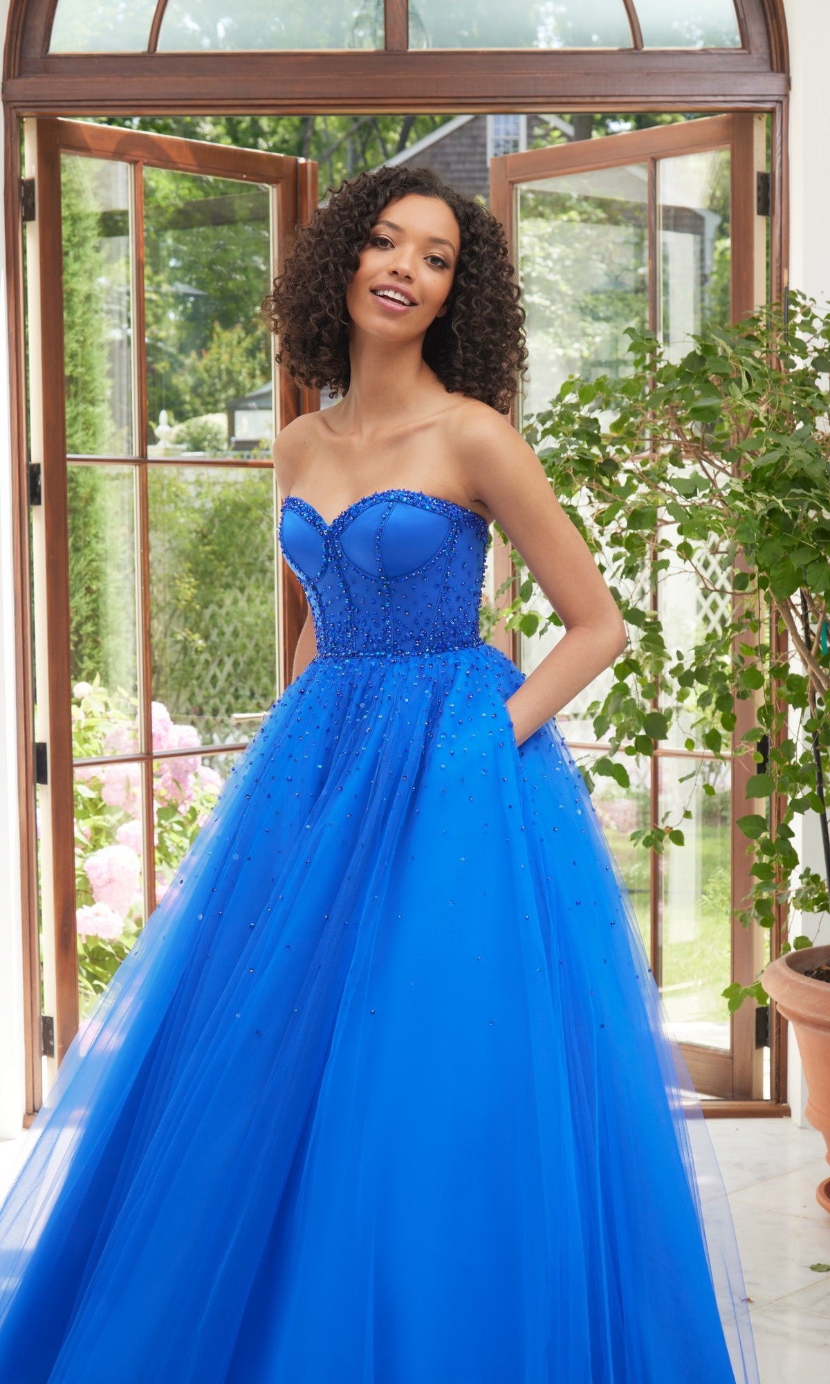 A woman stands indoors in the Morilee 49100 long prom dress, featuring a strapless sweetheart bodice, beaded details, and a tulle skirt in Regal Royal, framed by large windows and potted plants.