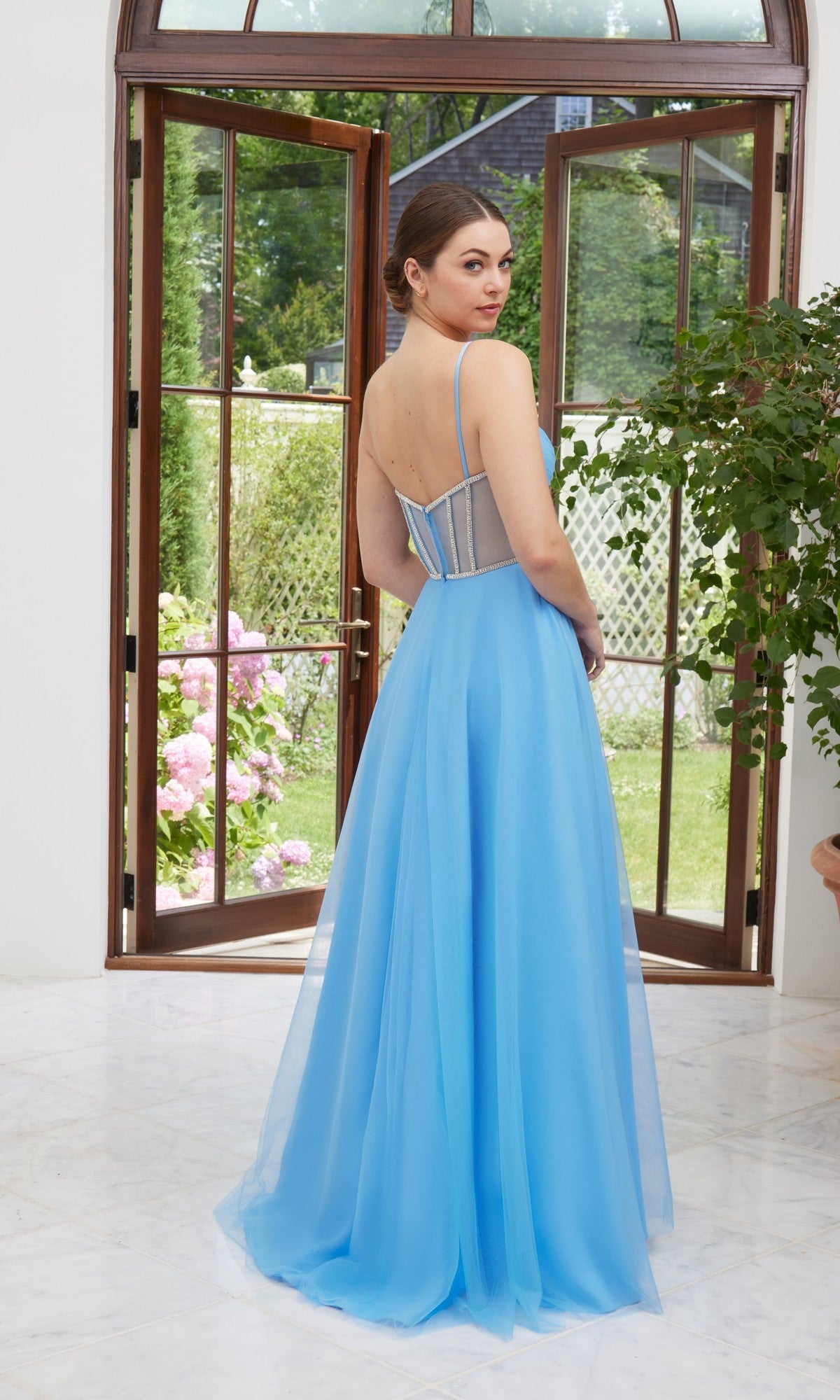 A woman stands indoors near open glass doors, wearing the Morilee 49114 long prom dress in French Blue. The gown features thin straps, a sheer back, and a corset bodice embellished with crystals as she looks over her shoulder.