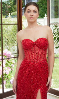 A woman stands indoors by large windows, wearing the Strapless Sequin Prom Dress: Morilee 49117 with a thigh-high slit. Greenery and pink flowers outside complement her sparkly dress.