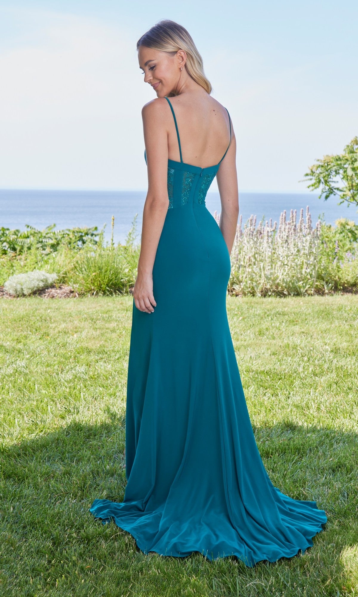 A woman stands on grass facing away, wearing the Long Prom Dress: Morilee 49127—a teal, floor-length gown with a lace bodice—set against a scenic outdoor backdrop with the ocean in the distance.