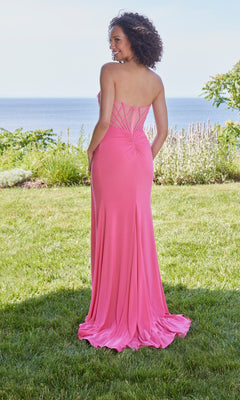 A woman stands on grass facing away, wearing the Morilee 49128 long prom dress. This strapless, floor-length pink gown features a ruched back and crisscross detailing, flowing elegantly with ocean and greenery behind her.