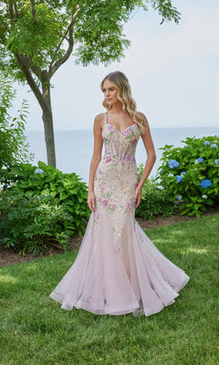 A woman stands on grass in front of bushes, wearing the Morilee 49130 long prom dress in blush/multi with a fitted bodice and sheer, floral-embellished skirt. Trees and water appear in the background.