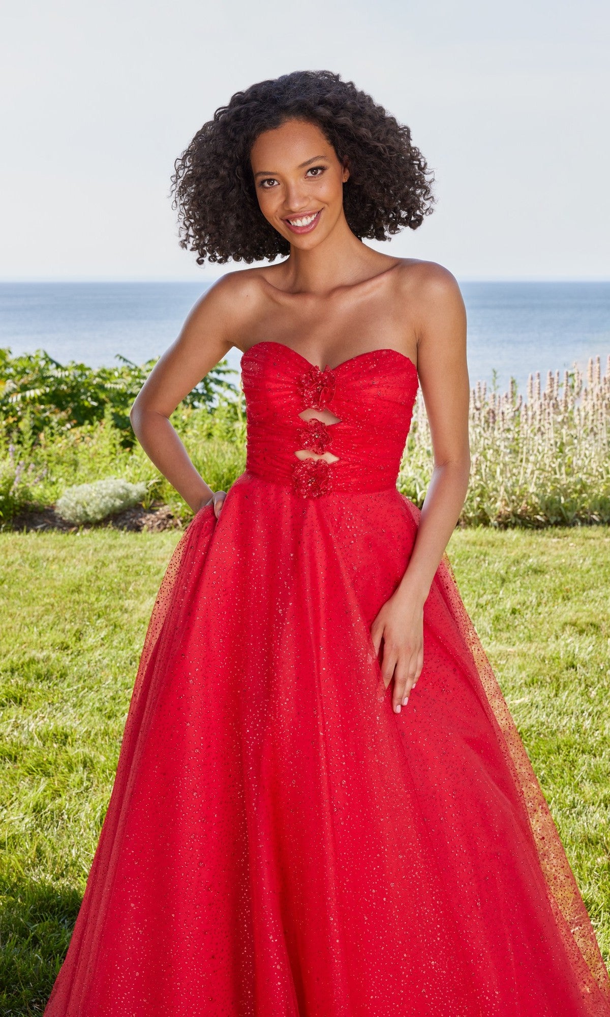 A woman in a scarlet Morilee 49137 long prom dress with an A-line skirt stands on grass, with shrubs and the ocean behind her.