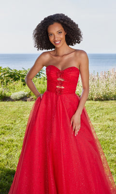 A woman in a scarlet Morilee 49137 long prom dress with an A-line skirt stands on grass, with shrubs and the ocean behind her.