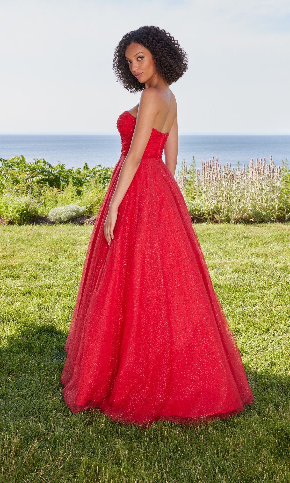 A woman stands on grass by the ocean and sky, wearing the Morilee 49137 long prom dress—a strapless ball gown with an elegant a-line skirt.