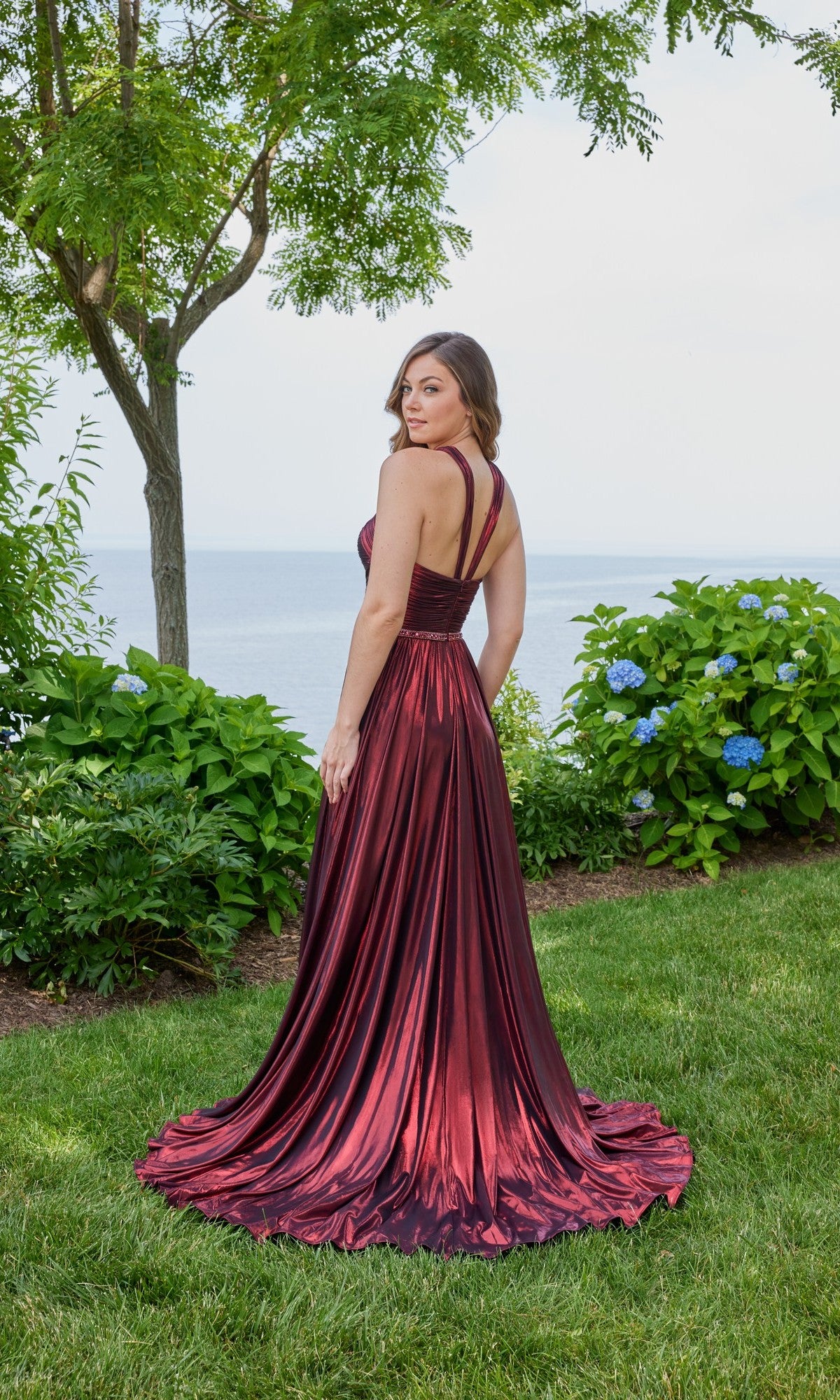 A woman wearing the Long Prom Dress: Morilee 49143 stands on grass, facing away from the camera, with trees, greenery, and water in the background.