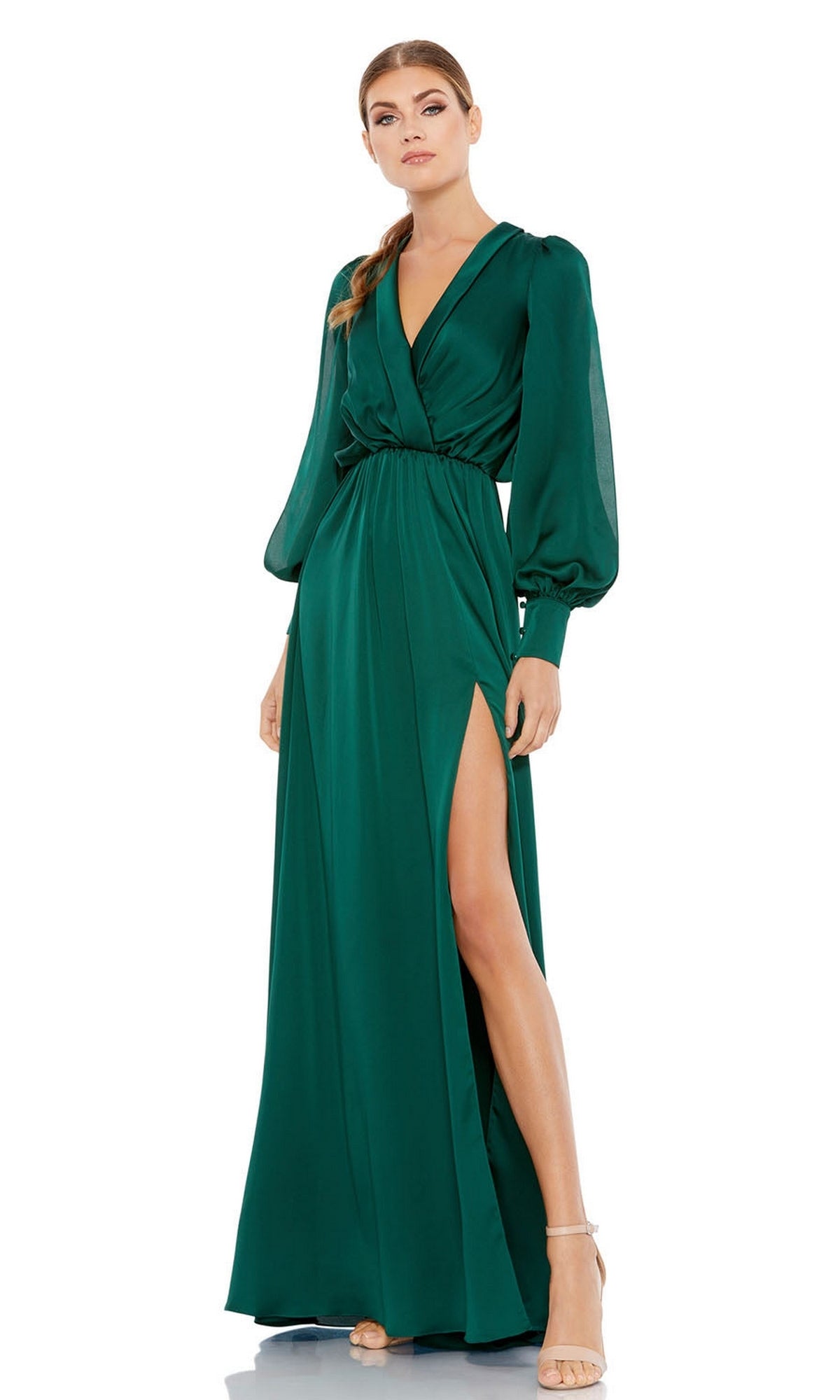 A woman models the Mac Duggal 49146, an emerald green long formal dress with long sleeves, a V-neckline, and a high slit. She pairs it with strappy nude heels and stands against a plain white background.