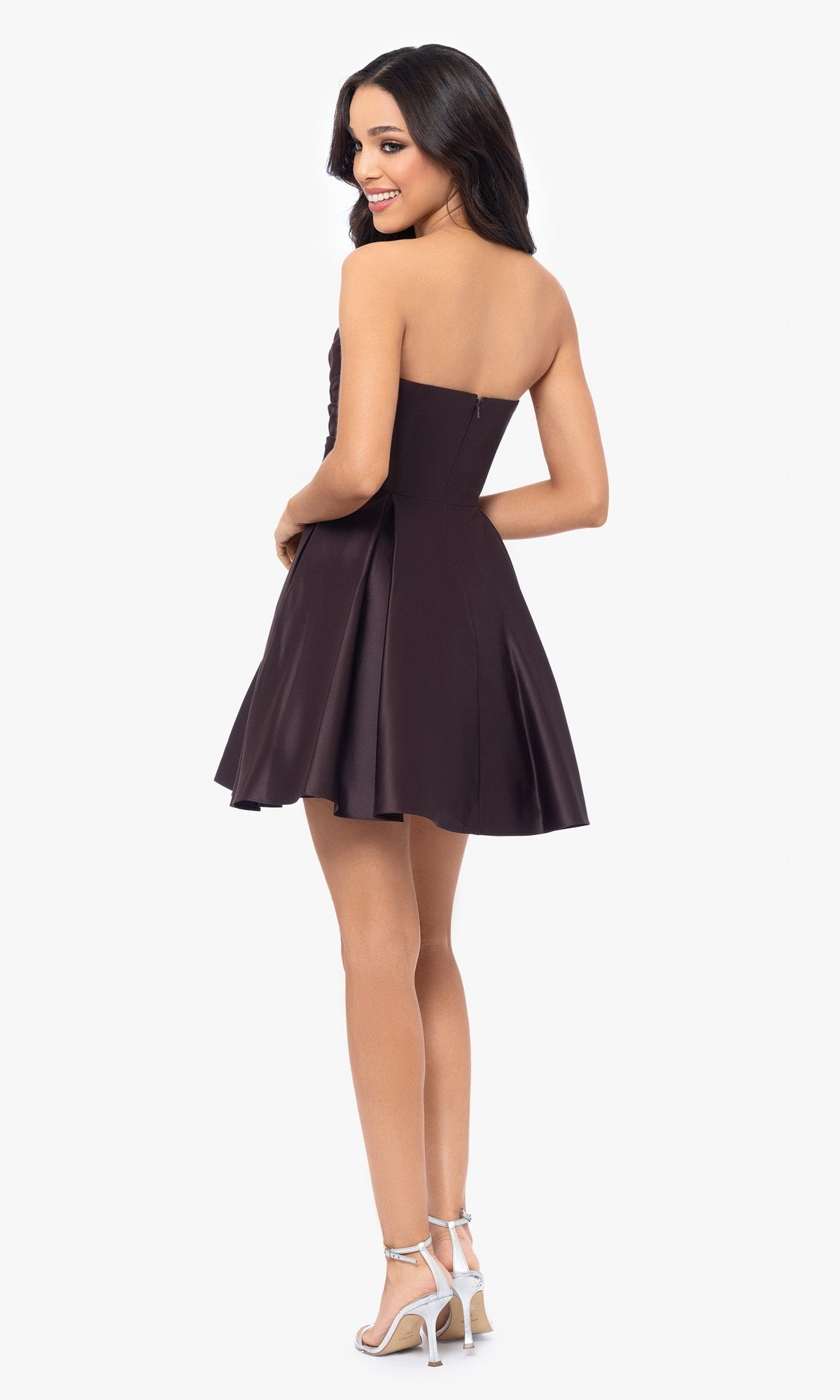 A woman, seen from behind, wears the Short Strapless Sweetheart Hoco Dress 4914BN with a ruched waist and silver high heels against a plain white background.
