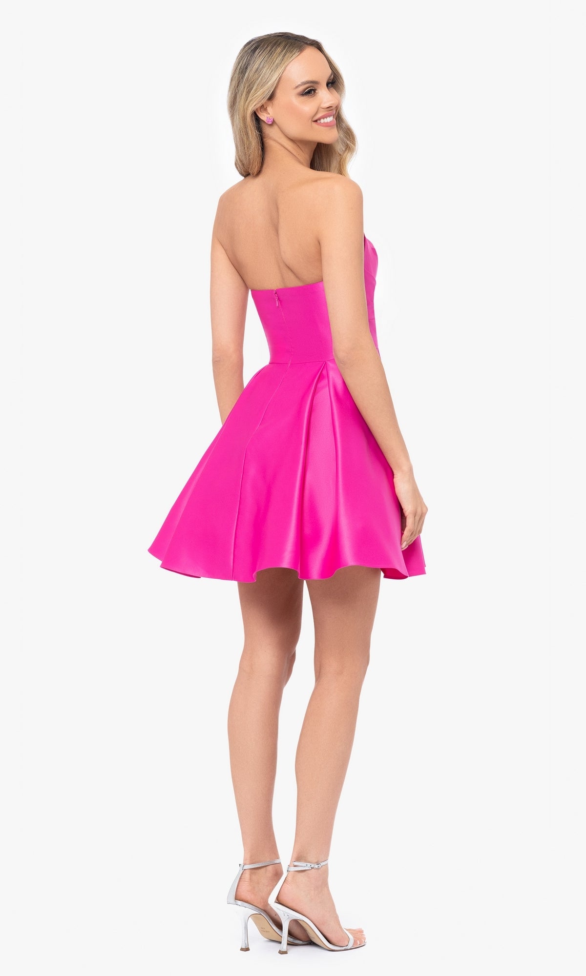 A woman with long blonde hair wears the Short Strapless Sweetheart Hoco Dress 4914BN in bright pink, featuring a ruched waist, paired with silver high-heeled sandals. She stands smiling over her shoulder.