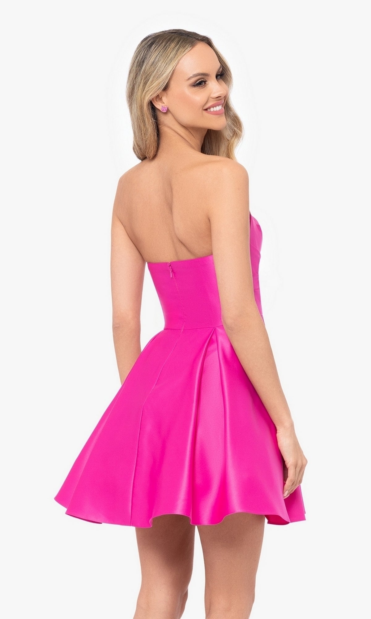 A woman wearing the Short Strapless Sweetheart Hoco Dress 4914BN in bright pink, featuring a fit-and-flare silhouette, stands facing away and smiles over her shoulder.