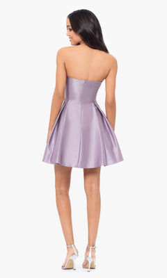 A woman models the Short Strapless Sweetheart Hoco Dress 4914BN, a knee-length lavender satin dress with a ruched waist, paired with silver high heel sandals, standing facing away from the camera against a plain white background.