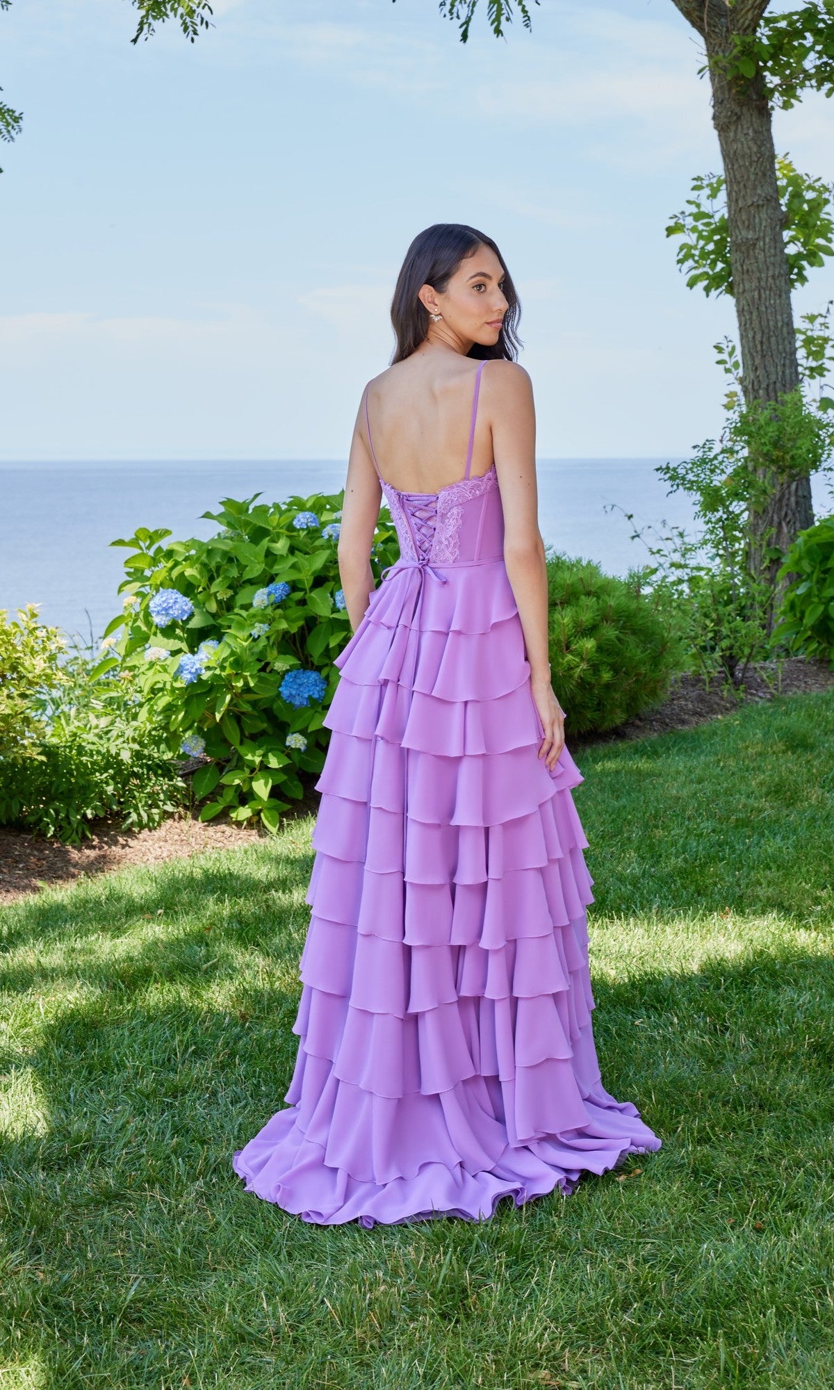 A woman stands on grass facing away, wearing the Morilee 49162 long prom dress in lavender with spaghetti straps and chiffon ruffles. Green bushes, trees, and water appear in the background.