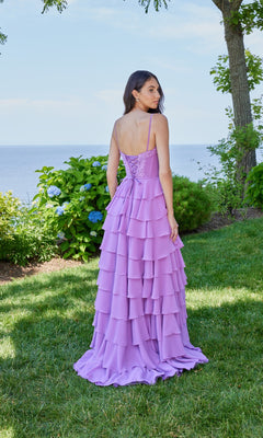 A woman stands on grass facing away, wearing the Morilee 49162 long prom dress in lavender with spaghetti straps and chiffon ruffles. Green bushes, trees, and water appear in the background.