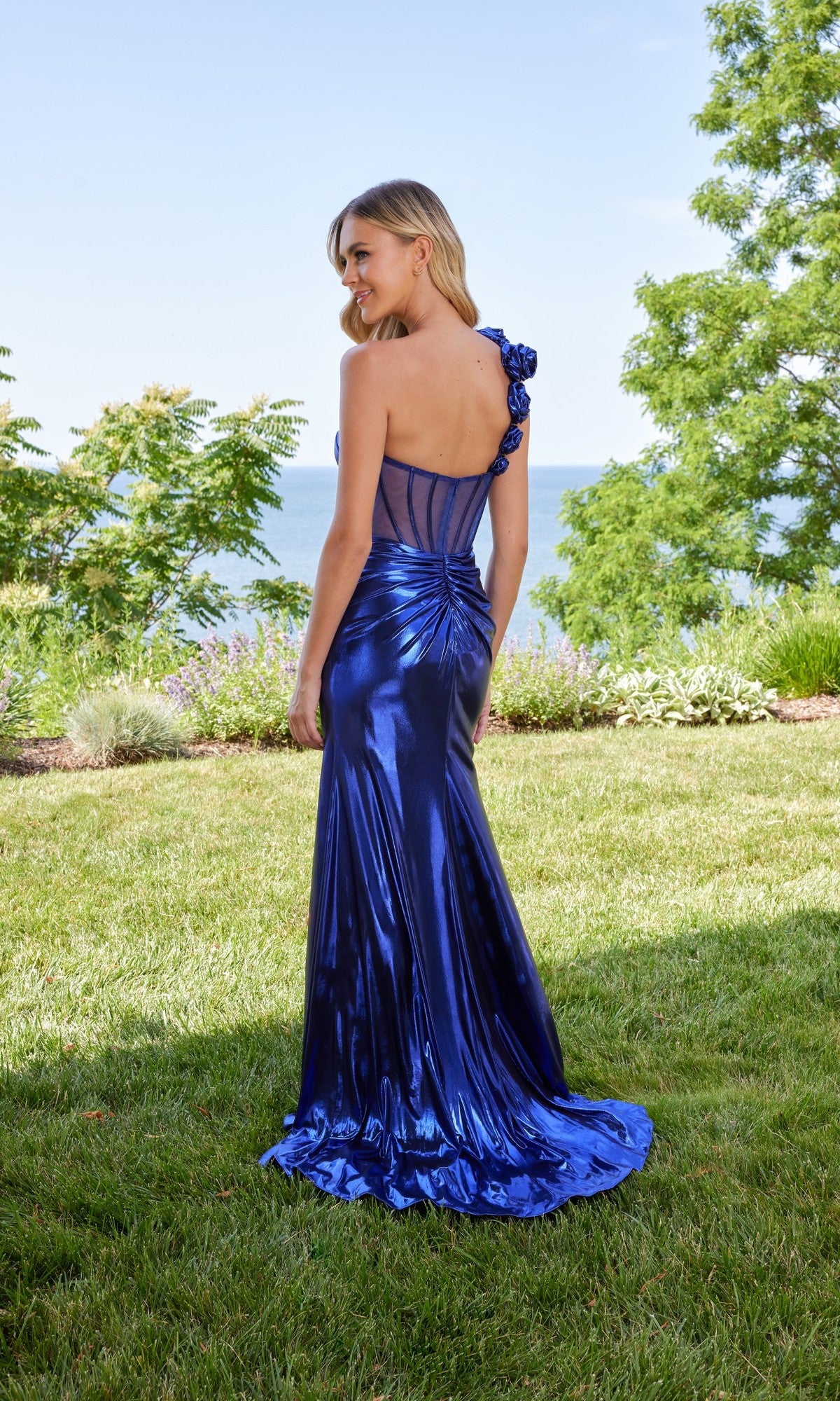 Wearing the Morilee 49165, a long metallic blue prom dress with an open back, a woman stands on grass facing away outdoors, with trees and water in the background.
