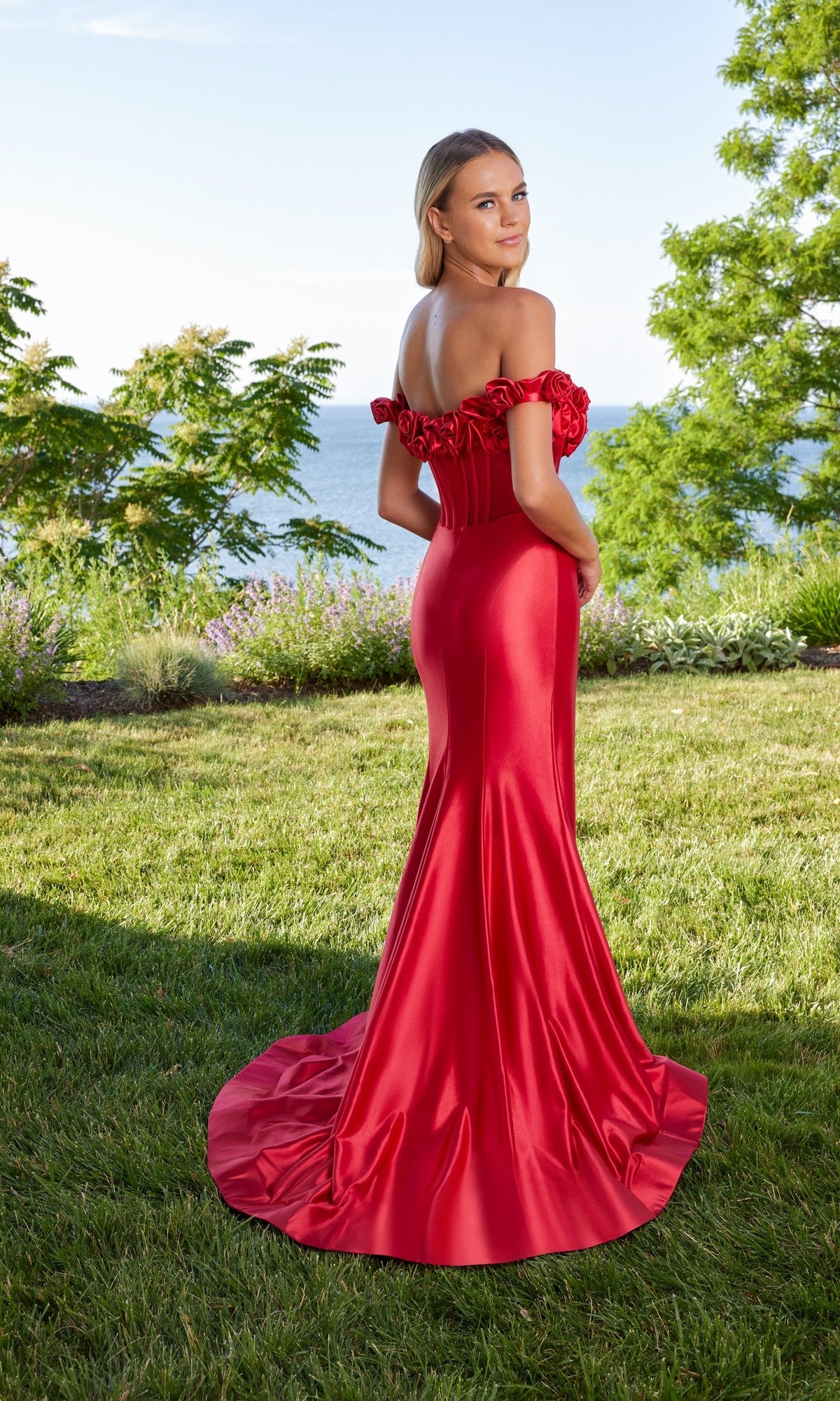 A woman wearing the Morilee 49171 long prom dress, fitted and off-the-shoulder, stands on grass facing away, with trees and water in the background.