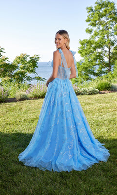 A woman stands on grass near water, wearing the Morilee 49185 long prom dress—a light-blue gown with silver patterns, an a-line tulle skirt, and shoulder bows. She smiles over her shoulder, with trees and shrubs behind her.