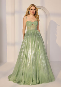 A woman wears the Morilee 49216 Long Prom Dress, a shimmery light green floor-length gown with a fitted corset bodice and thin straps, standing in a minimal, softly lit room.
